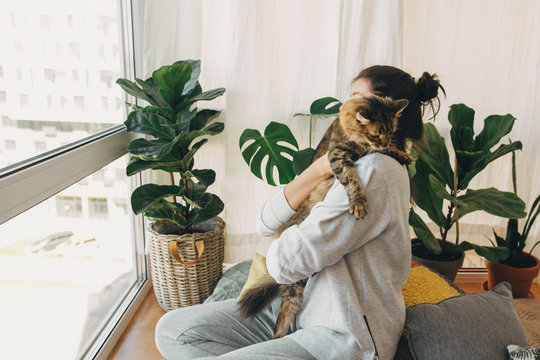 Hipster girl playing with cute cat, sitting together at home during coronavirus quarantine. Stay home stay safe. Isolation at home to prevent virus epidemic. Young woman with cat in modern room