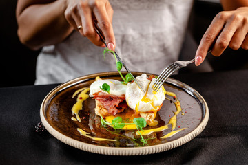 The girl in the restaurant has breakfast, toast with poached egg, salad, bacon and cheese sauce. Holding a knife and a fork in his hands. black background. copy space text