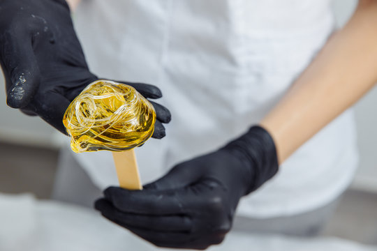 The wax is wound on a wooden spatula. The wax is intended for depilation. The spatula is held by hands in black latex gloves. Focus on the wax. The spatula is held by an epilation master.