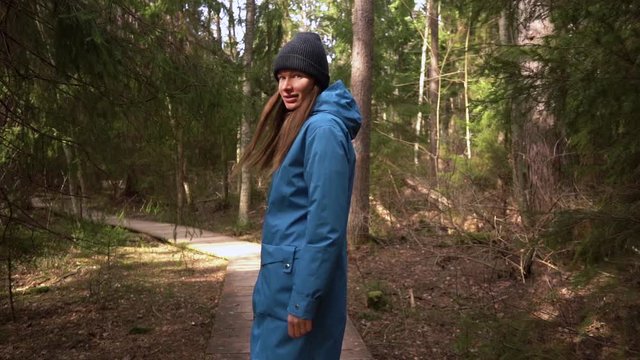 A woman in a blue raincoat and black hat is walking along an ecological trail and turns around and smiles. Slowmotion