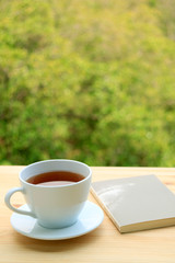 Vertical image of a cup of hot tea and a book on an outdoor table with blurry foliage in background