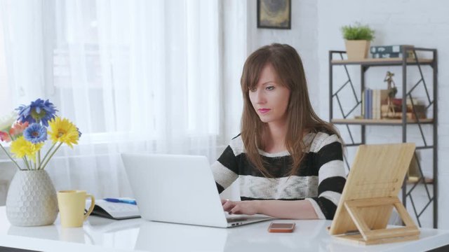 Woman working at desk with laptop computer at home office, thinking, looking busy. 4K video footage.
