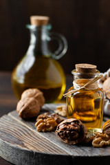 Concept of vegetable fat and oils for cooking and cosmetology. Glass bottle with bright yellow essence, raw walnut peeled and in their shells. Wooden board, dark wooden background, close up, macro