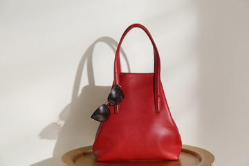 Stylish red woman's bag and sunglasses on table near light wall