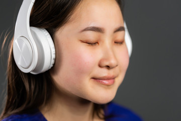 Young beautiful asian woman with headphones in modern wear enjoying and dancing on grey background. Radio, melody, technology concept.