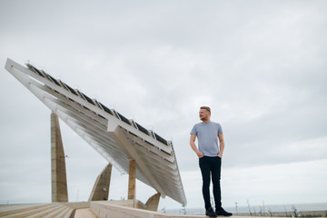 A young man in a t-shirt stands near big solar panel at Barcelona, Spain. Copy space