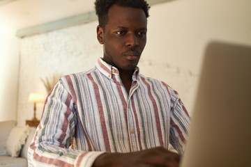 Serious concentrated young Afro American freelancer keyboarding on laptop working from home. Focused student learning online using electronic device, passing test. Technology, education and job