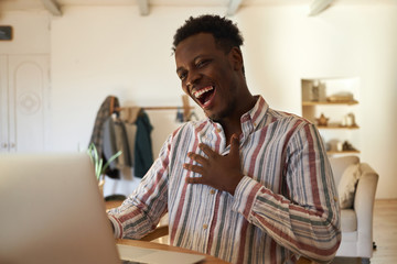 Cool charismatic young dark skinned man relaxing at home using laptop while surfing internet,...