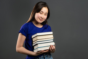 Asian student on grey background in studio holds stack of university books from library. Woman smiles, she is happy to graduate.