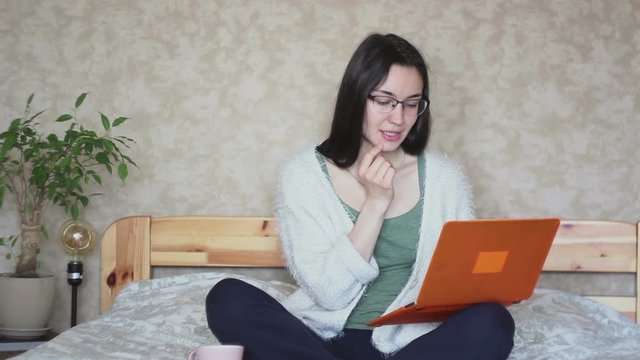Young attractive girl works remotely while sitting on the bed at home. Young Cute girl works at a laptop while sitting on the bed. Work at home.
