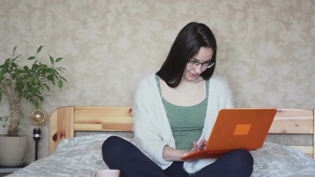 Young attractive girl works remotely while sitting on the bed at home. Young Cute girl works at a laptop while sitting on the bed. Work at home.