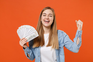 Joyful young woman girl in casual denim clothes posing isolated on orange background. People lifestyle concept. Mock up copy space. Hold fan of cash money in dollar banknotes, doing winner gesture.