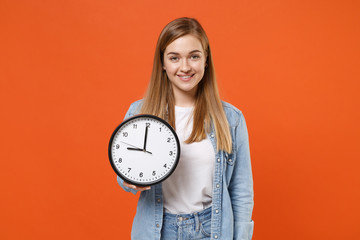 Smiling beautiful young woman girl in casual denim clothes isolated on bright orange wall background studio portrait. People sincere emotions lifestyle concept. Mock up copy space. Hold in hand clock.