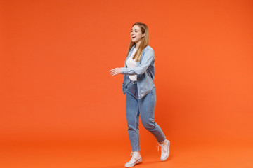 Fototapeta na wymiar Cheerful young woman girl in casual denim clothes posing isolated on orange wall background studio portrait. People lifestyle concept. Mock up copy space. Standing with outstretched hand for greeting.