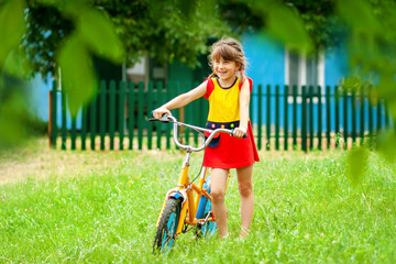 little girl in red dress stands on green grass next to an orange bicycle on background of green fence and blue village house. summer concept
