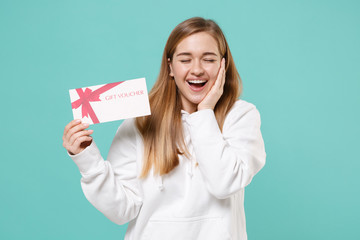 Funny young woman girl in casual white hoodie posing isolated on blue turquoise background. People lifestyle concept. Mock up copy space. Hold gift certificate, keeping eyes closed, put hand on cheek.