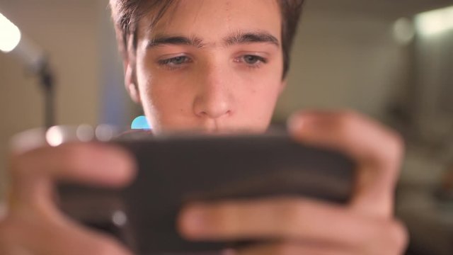 Close up face of a teenage boy with smartphone, at home. Caucasian teen guy spending time in a social network using mobile phone. White young man using cell phone for gaming.
