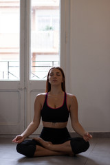View of young beautiful woman doing morning yoga after waking up at home. Female model sitting cross-legged in Easy pose, Sukhasana posture and meditating.