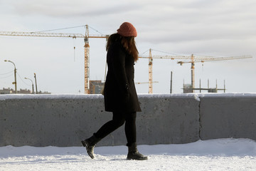 A girl in a hat and a black coat walks in the snow along a straight road against the background of...