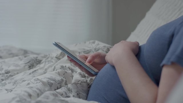 Pregnant Woman Relaxes In Bed, During Day, She Scrolls On Her Smartphone