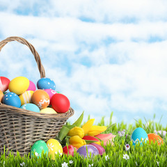 Wicker basket with Easter eggs in green grass against blue sky. Space for text