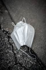 A close up of a dirty used white medical mask or surgical respiratory face mask discarded and laying as trash isolated on an asphalt street next to a concrete curb in the city.