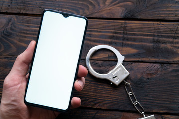 Man's hand hold a modern smartphone with a blank screen, handcuffs lie against a wooden background