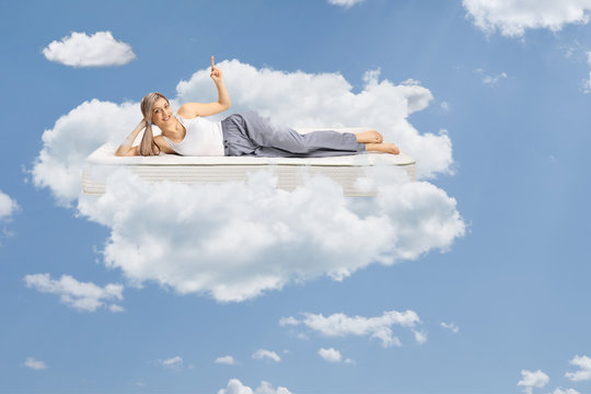 Young woman lying on a comfortbale mattress in pajamas, pointing up and floating on clouds
