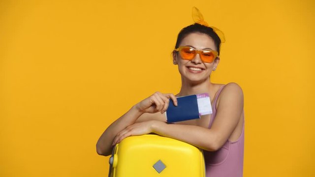 Smiling woman holding passport and air ticket near suitcase isolated on yellow