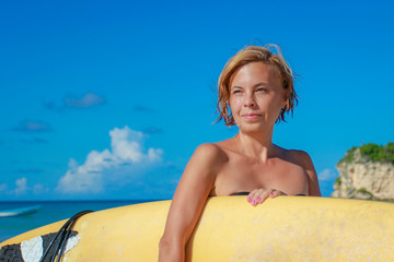 Surfer girl - novice on a tropical beach with a surfboard on a background of blue sky