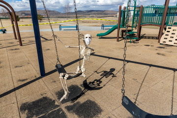 Skeleton swinging on childs toy wearing a mask at an empty closed park on a nice day