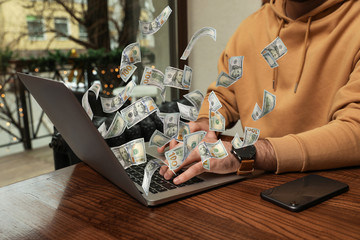 Man with modern laptop and flying dollar banknotes at table indoors, closeup. People make money online