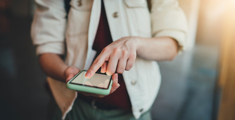 Frontal photo of young hipster girl in trendy white jeans jacket using modern smartphone typing sms message, closeup of female hands using cellphone with blank screen indoor, flare light effect