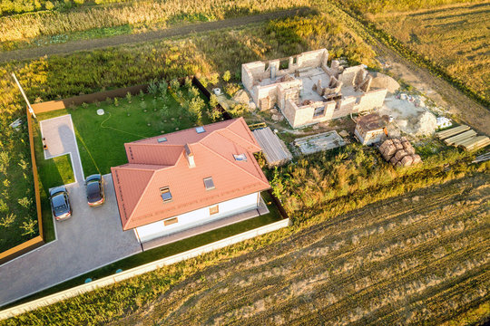 Top down aerial view of two private houses, one under construction with concrete foundament and brick walls and another finished with red tiled roof.