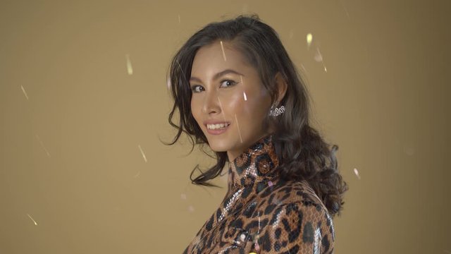 Asian modle poses for headshots with wind blowing her hair and sparkles and glitter in the wind on a beige background