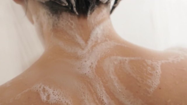 Naked woman takes a shower. Woman washes her short hair with shampoo. Slow motion video in white bathroom.