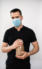 A man in a medical mask holds a jar of buckwheat on a white background.