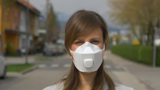 SLOW MOTION, CLOSE UP, PORTRAIT, DOF: Worried young woman wears a face mask while outdoors during covid19 quarantine. Woman squints while looking at camera as she stands outside with respiratory mask.