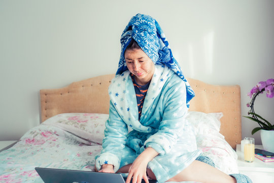 Portrait of a woman in a bathrobe and with a towel on her head, applying a scrubbing mask on her face while using a laptop, sitting on the bed. Lifestyle while working from home. Remote workplace.