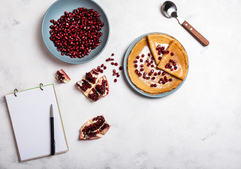 Grains of peeled pomegranate lie in a plate. Pancakes with pomegranate and condensed milk lie on a plate. View from above. Close-up. A book for writing recipes lies on the table. Copy space for text.