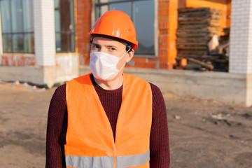Portrait of male construction worker in medical mask and overalls on background of house under construction.