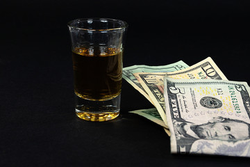 close up of folded American money and shot of whiskey on black