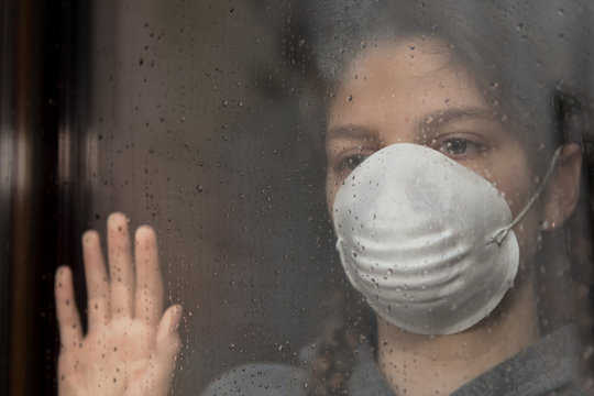 COVID-19 Pandemic Coronavirus, Young girl in home isolation, during quarantine is wearing protective face mask for stave off preading of disease virus, looking out of the window