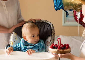 One year old cute boy and berries cake with a candle. Boy wearing celebration shirt, sitting on a highchair. First Happy Birthday at home. Family concept.