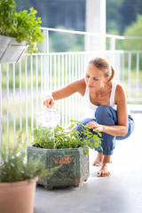 Pretty, young woman watering herbs she is growing on her balcony.