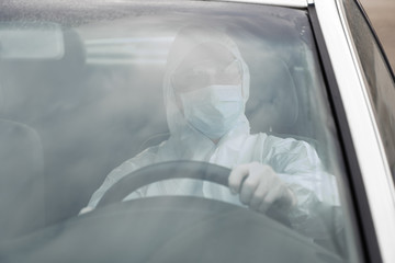 Man in protective suit, medical mask and rubber gloves for protect from bacteria and virus is driving a car. Protective mask while quarantine, world pandemic, covid 19, coronavirus, infection.