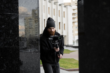 Fototapeta na wymiar Bearded Professional videographer in black hoodie holding professional camera on 3-axis gimbal stabilizer. Filmmaker making a great video with a professional cinema camera. Cinematographer.