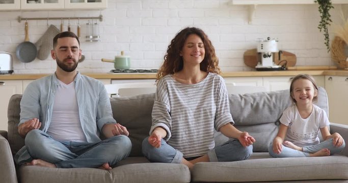 Healthy calm funny family mom dad and little cute daughter meditate together with eyes closed sit on sofa, mindful young parents teach child girl do yoga exercise relax together in lotus pose at home