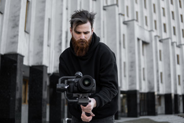 Bearded Professional videographer in black hoodie holding professional camera on 3-axis gimbal stabilizer. Filmmaker making a great video with a professional cinema camera. Cinematographer.