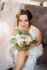 A serious bride with a bouquet in her hands sits on a soft chair in the room.